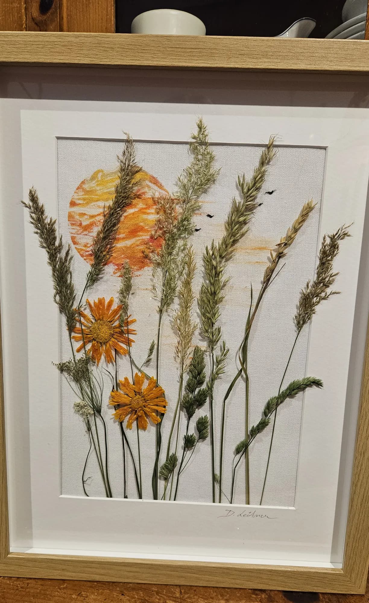 Gerahmtes Bild mit gemalter Sonne, orangen Blüten und getrockneten Gräsern auf Leinen