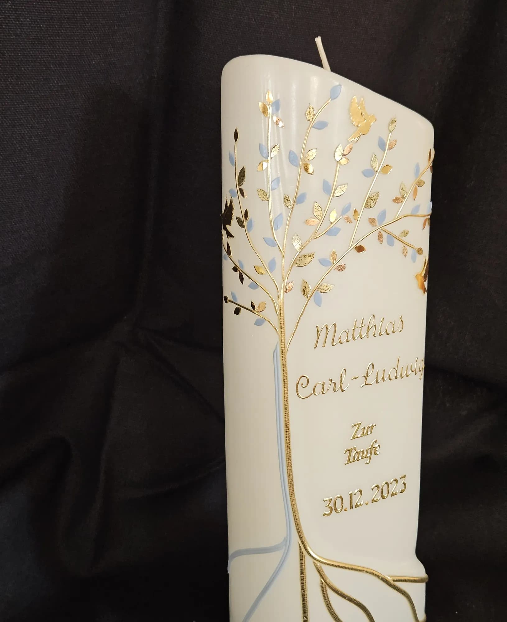 Cremefarbene Taufkerze mit goldenem Lebensbaum vor dunklem Hintergrund