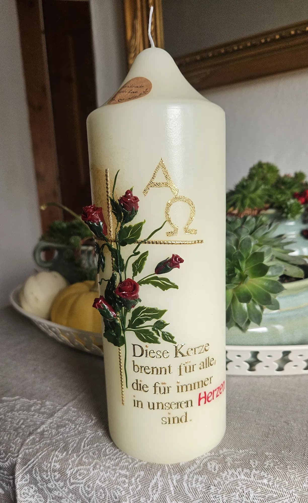 Cremefarbene Trauerkerze mit roten Rosen, goldenem Kreuz sowie Alpha und Omega